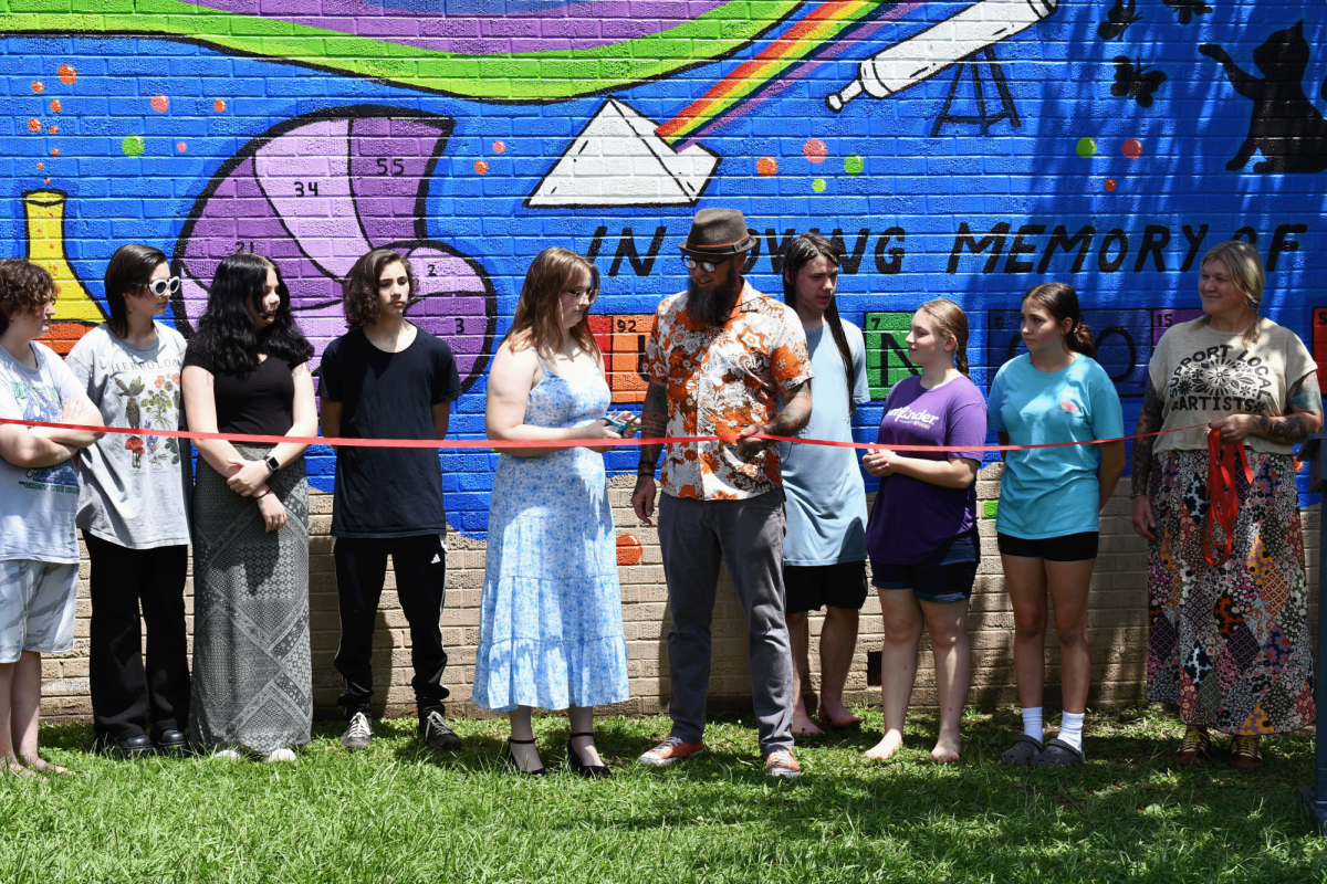 A girl in a blue dress cuts a red ribbon with support from a man in a floral shirt as others stand beside them in front of a colorful science-themed mural.