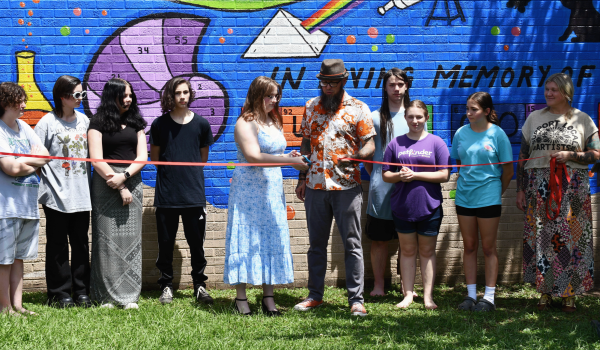 A group of students and adults stand in front of a blue mural with science-themed images and the words “In Loving Memory of” as a girl in a blue dress holds ribbon-cutting scissors.