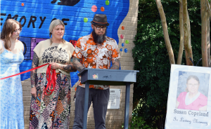 A man in a floral shirt and hat speaks at a podium in front of a colorful mural as two women stand beside him holding red ribbon. A memorial sign with a photo of Susan Copeland is displayed to the right.