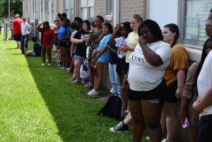 A group of students stands along a school building wall on a grassy lawn, watching the mural unveiling event.