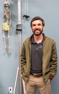Physical therapist Kollin Cannon smiles while standing beside a skeleton model in a treatment room.