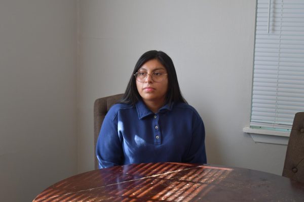 A young woman with long dark hair and glasses sits at a table near a window. She is wearing a blue collared shirt and looking slightly to the side.