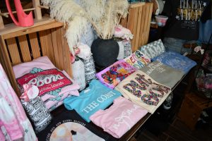 Display table inside The Charm Bar boutique in Wiggins, Mississippi, with folded T-shirts, flowers, and accessories arranged beneath a colorful checker-patterned sign.