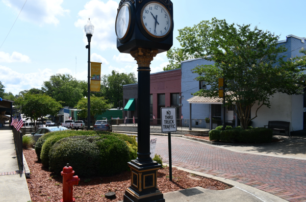 Black street clock with gold trim stands beside a red fire hydrant and a “No Thru Truck Traffic” sign in downtown Wiggins, Mississippi. Brick roads and storefronts line the street.