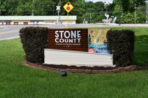 Sign reading “Stone County Economic Development Partnership” with a colorful mosaic sits beside a rural road and railroad tracks in Wiggins, Mississippi.