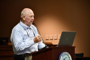 Joe Spraggins speaks at a podium with a laptop and microphone during an MDMR public meeting.
