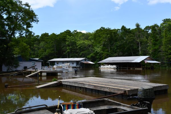 Wooden dock and small boats sit along a calm riverbank with multiple houseboats and cabins in the background, surrounded by forest.