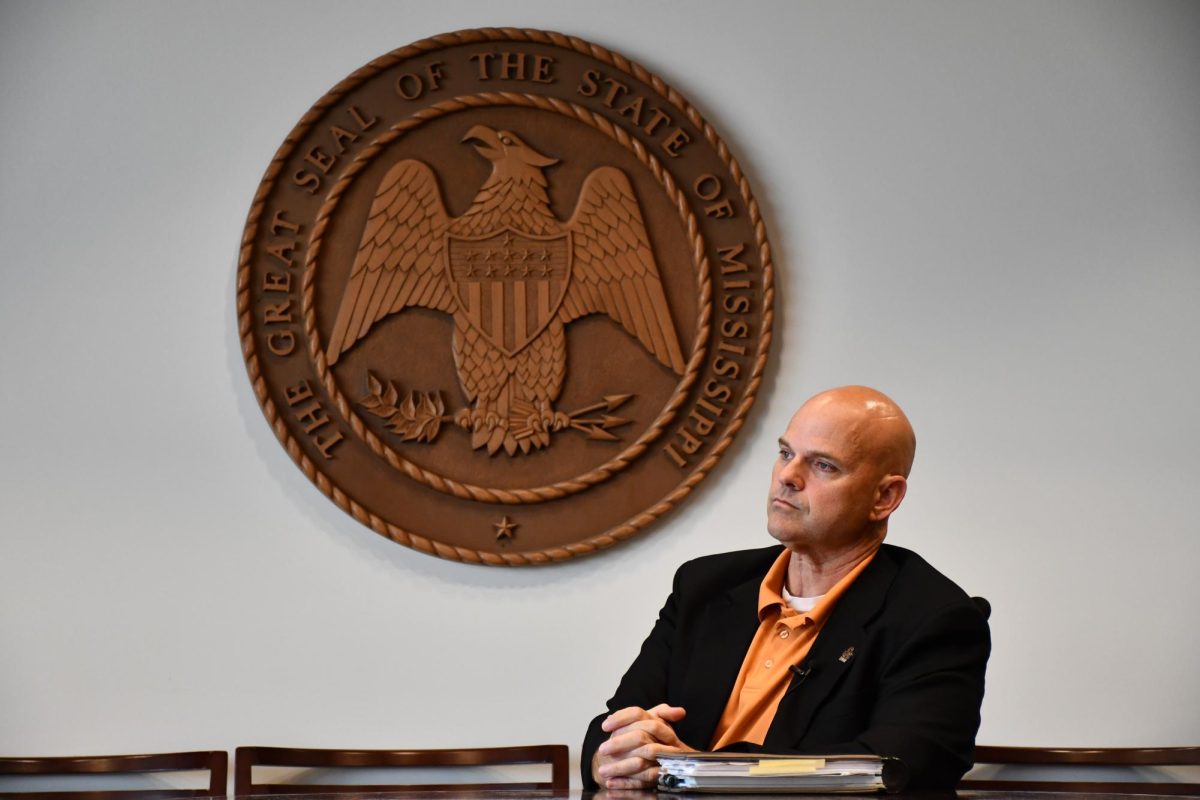 Man in a black blazer sits beneath the Great Seal of the State of Mississippi.