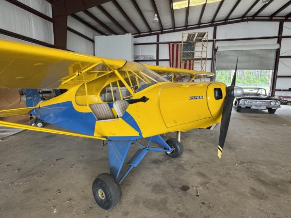 A yellow and blue single-engine CubCrafters airplane parked inside a large metal hangar with file cabinets and chairs along the walls.