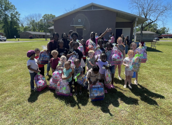 Children and volunteers pose outside on a sunny day, holding up colorful Easter baskets wrapped in plastic and filled with candy and toys.
