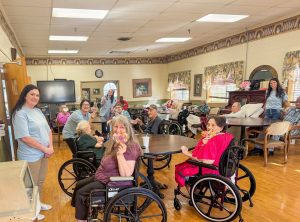 Junior Auxiliary volunteers smile alongside elderly residents in wheelchairs during a group activity in a care facility’s common room.