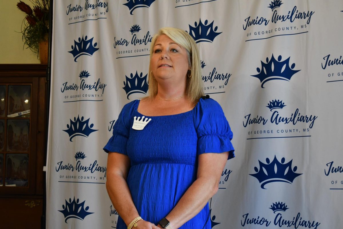 Gena Fortenberry, president of the Junior Auxiliary of George County, stands in front of a branded backdrop, speaking during a membership event.
