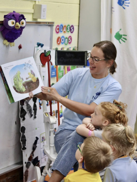 A woman reads a children’s book to a small group of young students gathered closely around her in a colorful classroom.