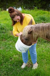 Trannie Tucker, wearing a yellow shirt and blue jeans, feeds a horse from a white bucket in a grassy field near a water source.