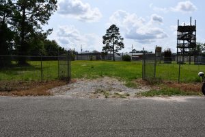 Road in front of fenced grassy area with gravel entrance and black chain link fence around the outside.
