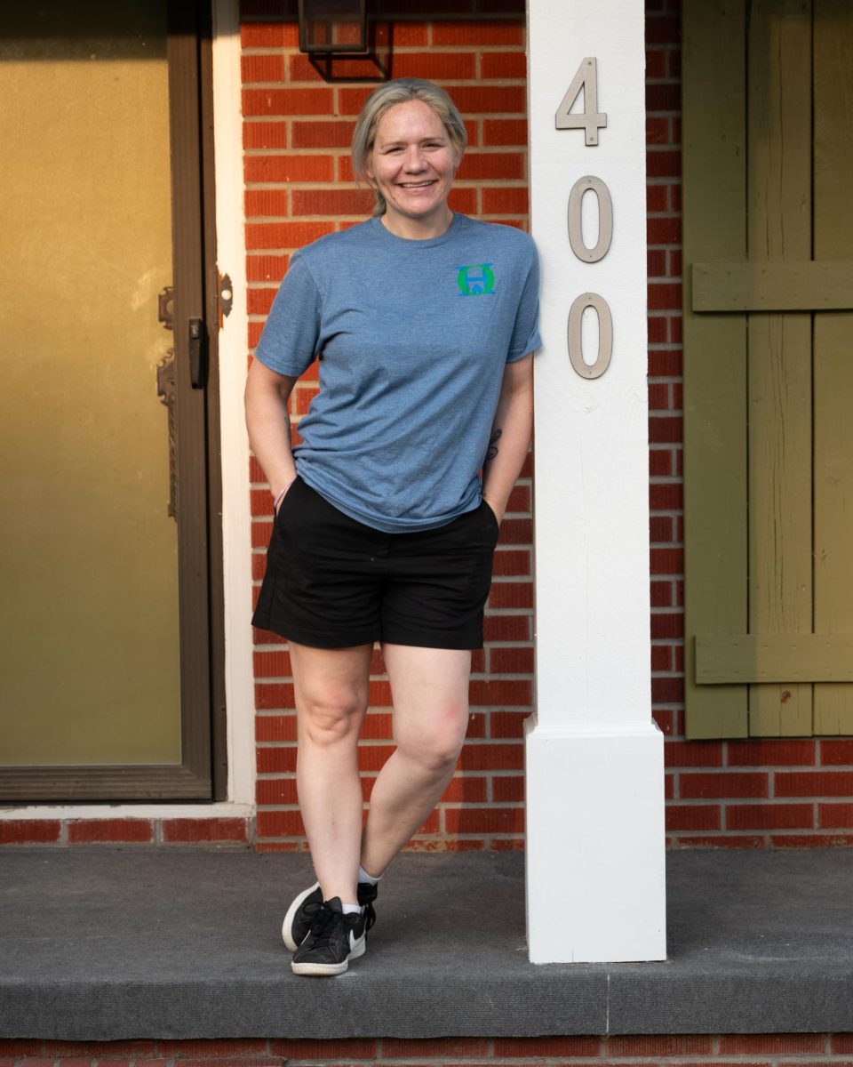 A woman stands on the front porch of a red brick house beside a white column labeled “400,” smiling and wearing a blue T-shirt and black shorts.