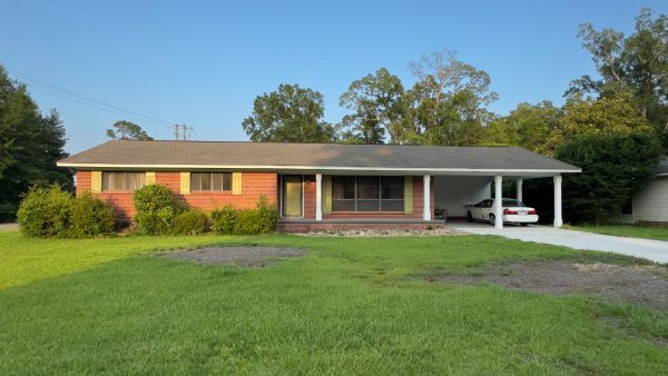 A single-story red brick house with a covered carport and a white car parked in the driveway, surrounded by green grass and trees.
