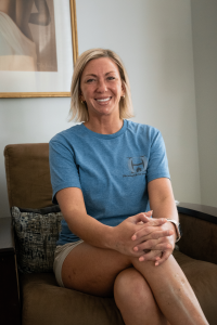 A woman smiles while seated in an armchair inside a well-lit room, wearing a blue T-shirt that reads “Strength in numbers.”