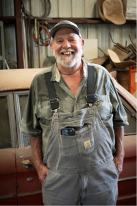 Richard Andersen in light blue Carhartt’s men’s bib overall with a light green shirt underneath stands inside a workshop in front of an old dusty red car.