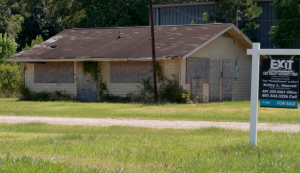 Abandoned building with broken roof and boarded windows stands next to a for sale sign in grassy lot.