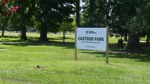 Sign for Eastside Park stands in grassy area surrounded by trees and walking paths.