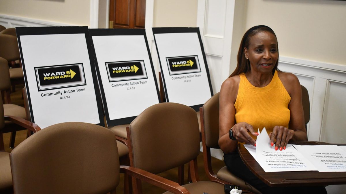 Woman in yellow top speaks while seated beside signs for the Ward 5 Community Action Team.