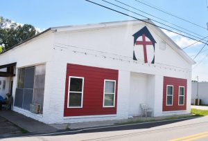 A single-story white brick building with red window panels and a red cross emblem above the front door. The building sits along a roadside under clear skies.