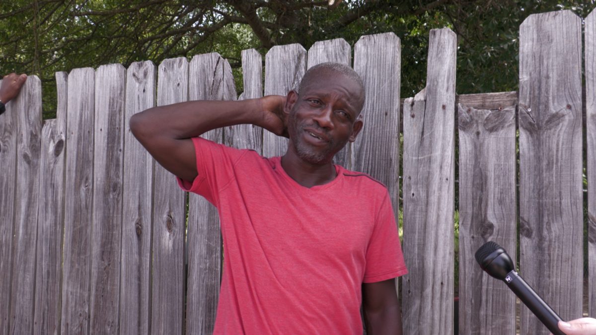A man in a red T-shirt stands in front of a wooden fence with one hand behind his neck, looking toward a microphone held by an interviewer.
