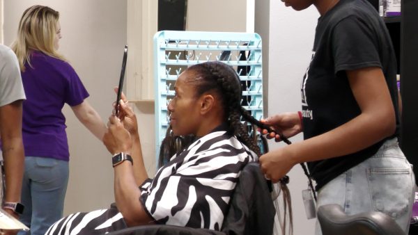 A cosmetology student braids a client's hair while the client looks into a handheld mirror during a training session at a beauty school.