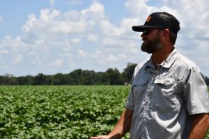 Farmer wearing sunglasses and a cap smiles while standing in front of a cotton field.