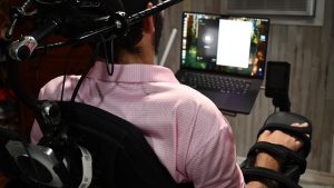 A man in a wheelchair, seen from behind, faces a laptop as he uses a Neuralink interface mounted on his chair in a home setting.