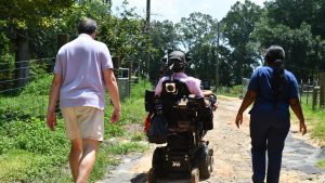 A man in a motorized wheelchair rides between two people on a rural path lined with trees and fencing.