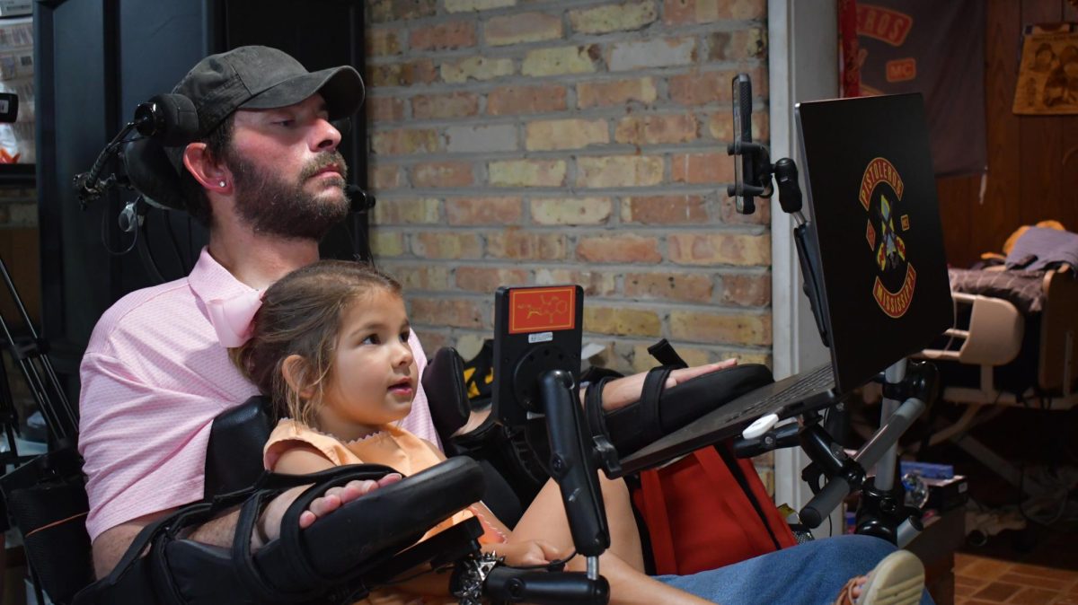 A man in a wheelchair sits with a young girl on his lap, both looking at a laptop screen.