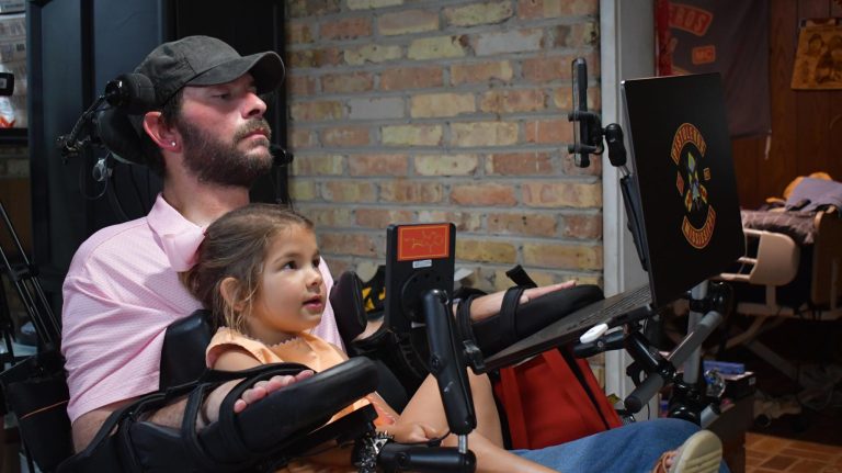 A man in a wheelchair sits with a young girl on his lap, both looking at a laptop screen.