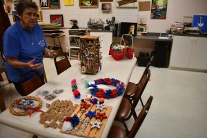 Senior woman in blue shirt prepares red, white and blue wreaths, earrings and pom-pom crafts at a long table covered in handmade items.