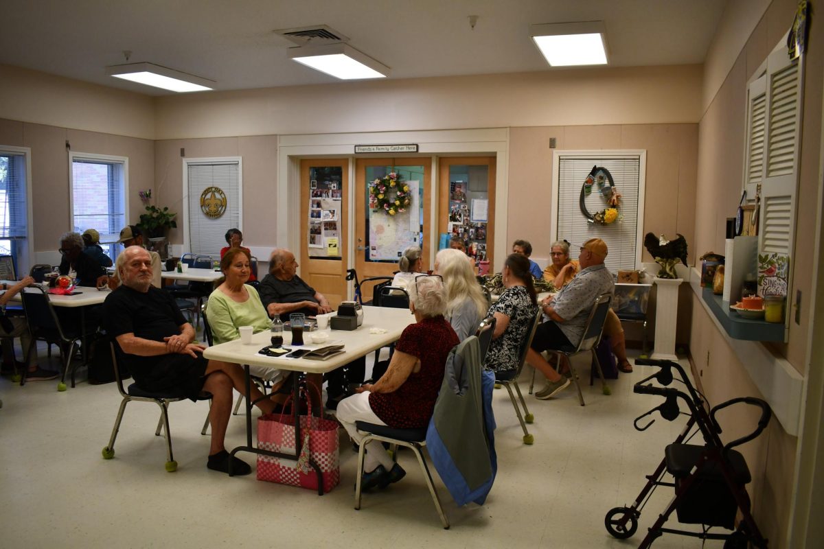 Group of older adults sit around tables in a community room decorated with photos and wreaths.