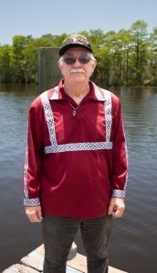 An older man wearing a decorated maroon shirt and military cap stands on a wooden dock near a body of water.