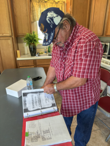An older man in a red plaid shirt and baseball cap looks through a large binder filled with printed documents on a kitchen counter.
