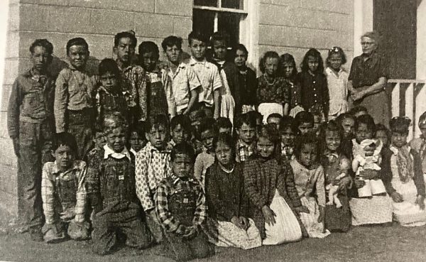 Black-and-white photo of dozens of school-age children and one adult standing and kneeling in rows outside a cinder block building.