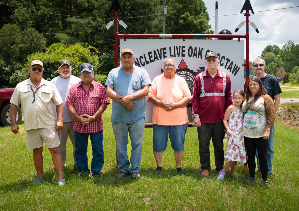 Nine people stand on grass in front of a large tribal sign, some with arms folded, others with hands at their sides.