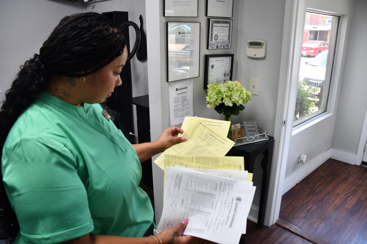 Asheena Woods, wearing a green outfit, looks down at several pages of yellow and white inspection and licensing forms inside her barber school office.
