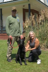 A man and a woman pose with a black German shepherd on a leash in front of a house. The man, wearing a green long-sleeve shirt, cap and sunglasses, holds the leash while the woman, dressed in black athletic wear, kneels beside the dog and smiles at the camera.