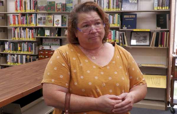 Woman wearing glasses and a yellow floral shirt stands in a library, speaking about the need for food pantries.