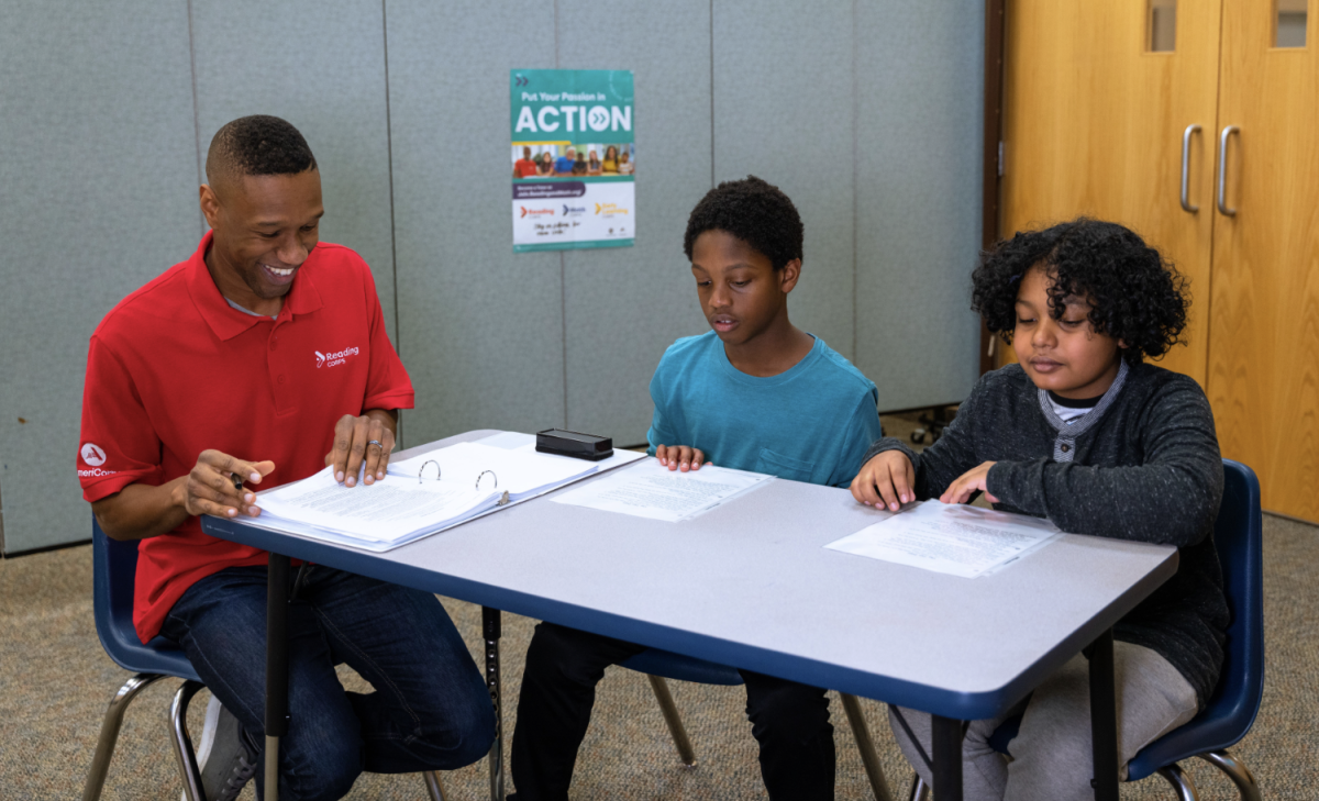 Tutor sitting at a table with two elementary students, reading and reviewing materials together.
