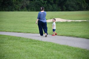 A woman walks on a paved track in the park while her young son, wearing red shoes, looks back toward the camera.