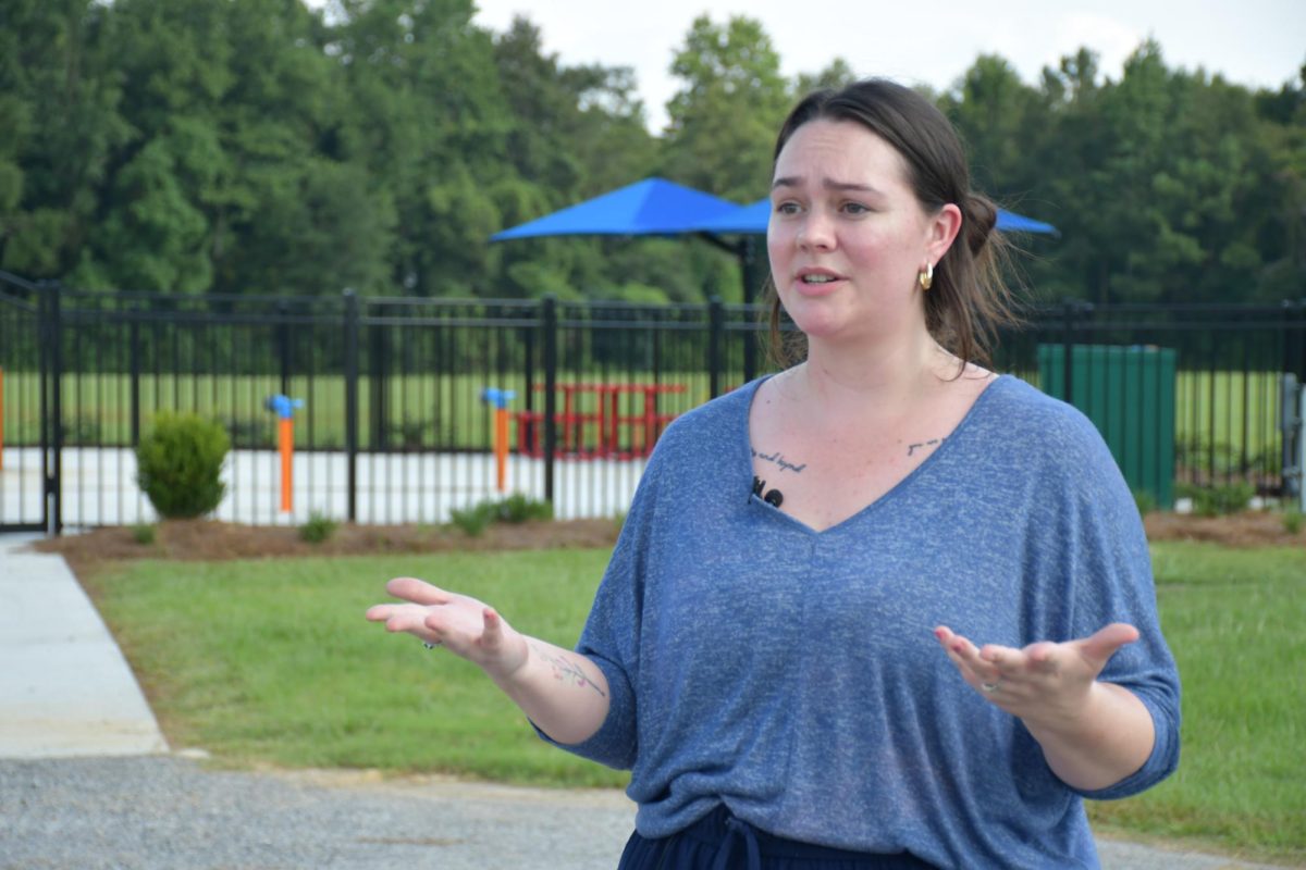 A woman in a blue shirt gestures with open hands while standing near Richton’s fenced splash pad.