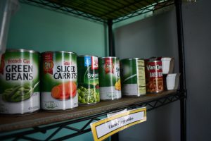 Cans of vegetables, like beans and corn, line a shelf inside a small food pantry.