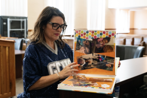 A woman wearing glasses turns pages in a colorful scrapbook inside a courtroom.