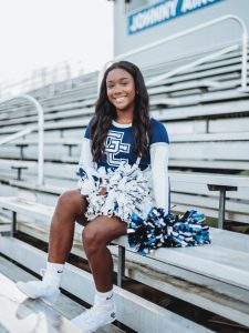 A smiling teenage girl wearing a blue cheerleading uniform sits on stadium bleachers holding silver pom-poms.