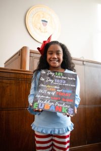 A young girl stands in a courtroom holding a colorful sign marking the day of her adoption.